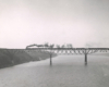 Black-and-white photo of steam locomotive crossing river with freight train on deck-truss bridge