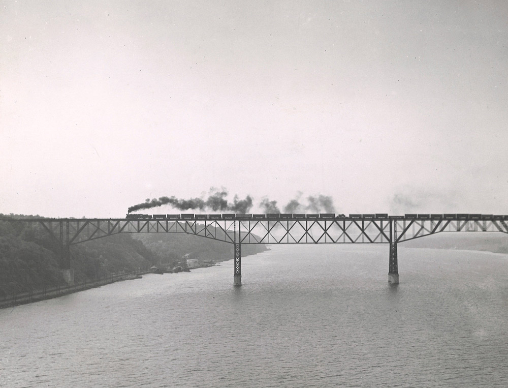 Black-and-white photo of steam locomotive crossing river with freight train on deck-truss bridge