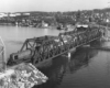 Black-and-white overhead photo of road-switcher diesel locomotive crossing through-truss bridge with freight train