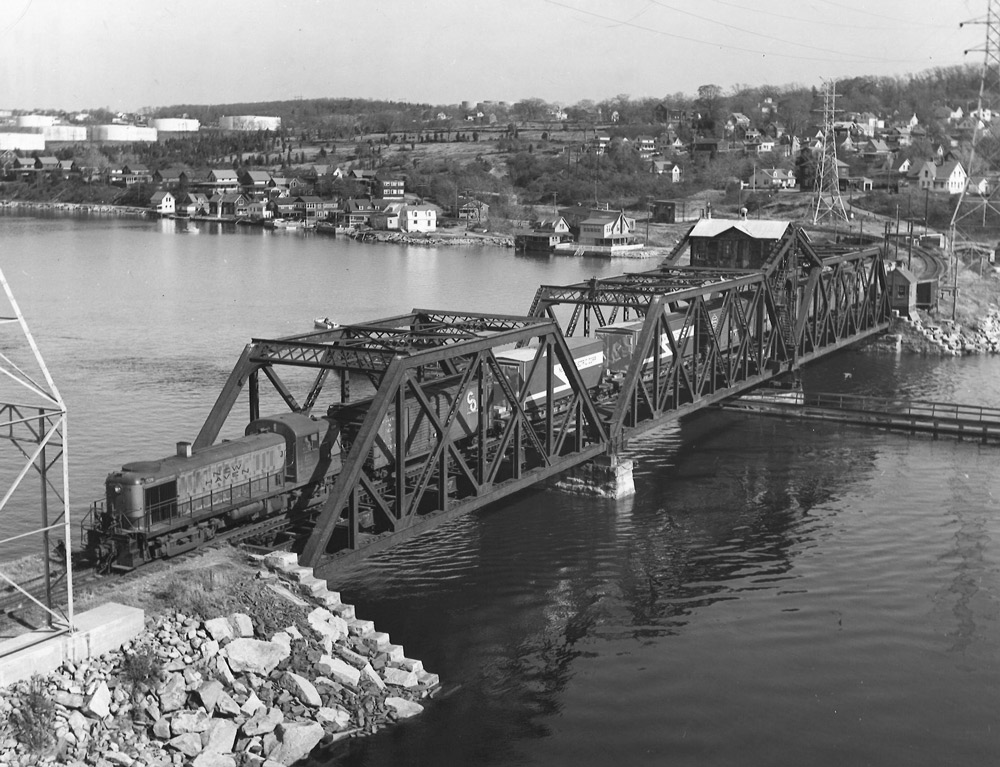 Black-and-white overhead photo of road-switcher diesel locomotive crossing through-truss bridge with freight train