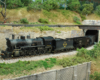 A steam locomotive leads a train out of a tunnel on the side of a wooded hill