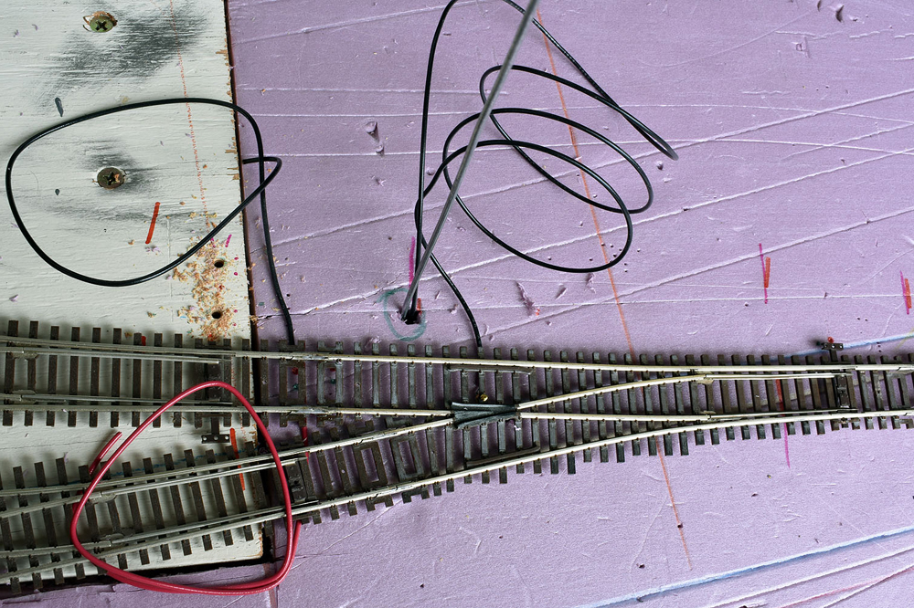 Three turnouts with wires attached, baling wire pushing black wire into hole in pink extruded-foam insulation board layout surface