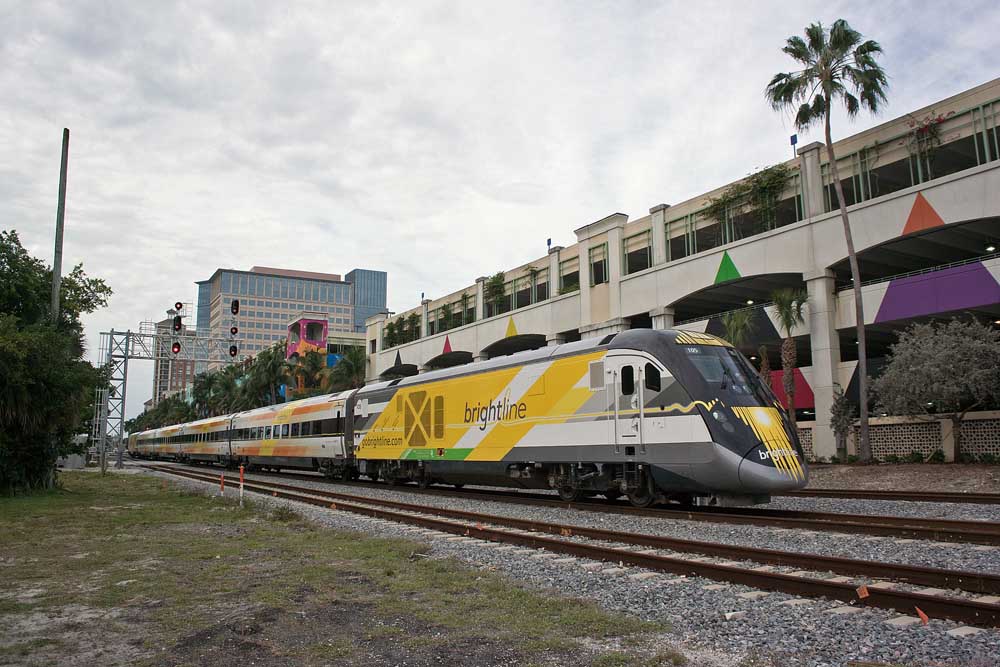 Multi-colored streamlined passenger train passes colorful parking garage and palm trees.