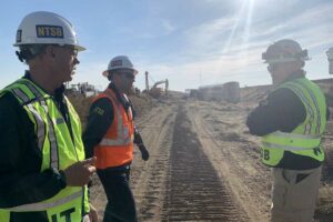 Three men in hard hats and safety vests in front of derailed passenger train