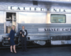 Blonde woman in dress and man in suit standing next to passenger car on station platform.