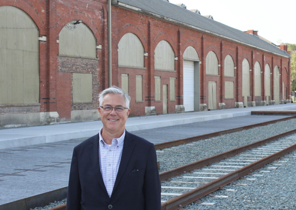 Man in blue sportcoat stands in front of brick factory building