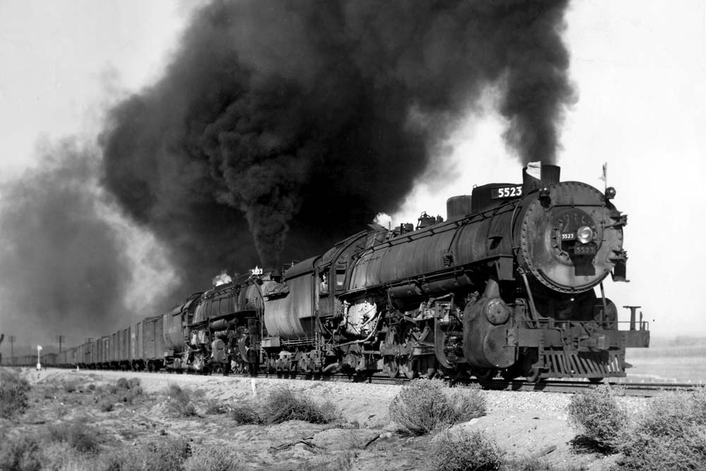 Two steam locomotives lead a freight train under heavy smoke