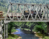 A geared steam locomotive crosses a wood truss bridge over a creek in a wooded scene