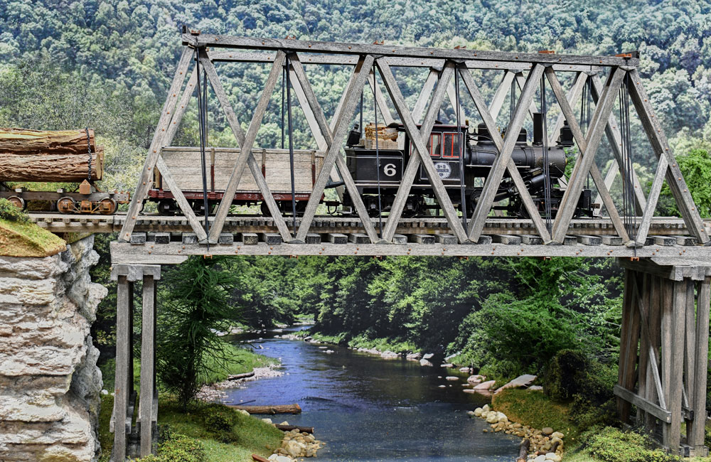A geared steam locomotive crosses a wood truss bridge over a creek in a wooded scene
