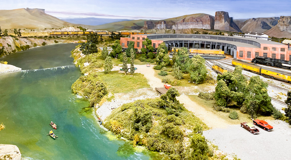 A broad, green river flows past a roundhouse and locomotive servicing terminal where yellow-and-gray locomotives ride the turntable