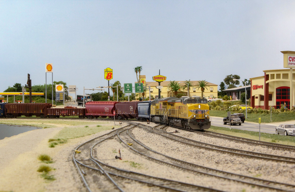 A modern yellow-and-gray diesel pulls a manifest freight past a busy highway lined with businesses