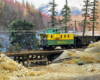 Railfans ride a green-and-yellow caboose behind a coal train as it crosses a short wooden trestle over a creek