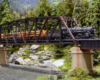 A steam locomotive pulls a string of loaded log cars over a steel trestle above a creek in a forest