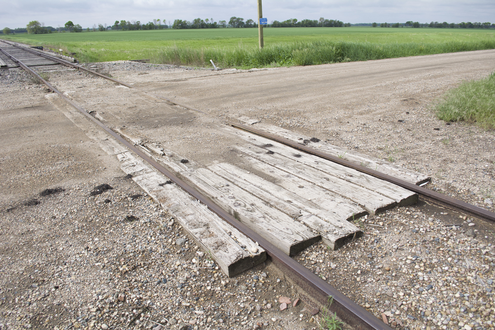 Gravel covered and worn wood planks at a rural grade crossing with a farm field in the background.