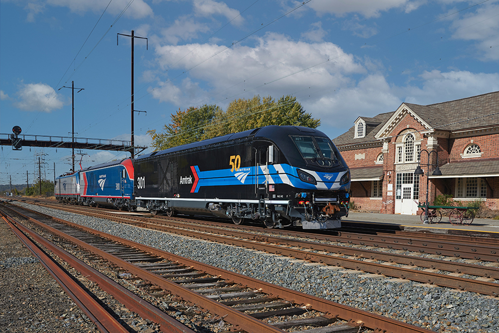 Two diesels — black locomotive with blue stripe and blue and red locomotive — pulled past station by electric locomotive