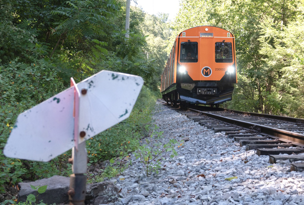 Orange railcar approaches switch on rail line surrounded by trees