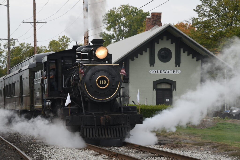 Small black steam locomotive departs train station.