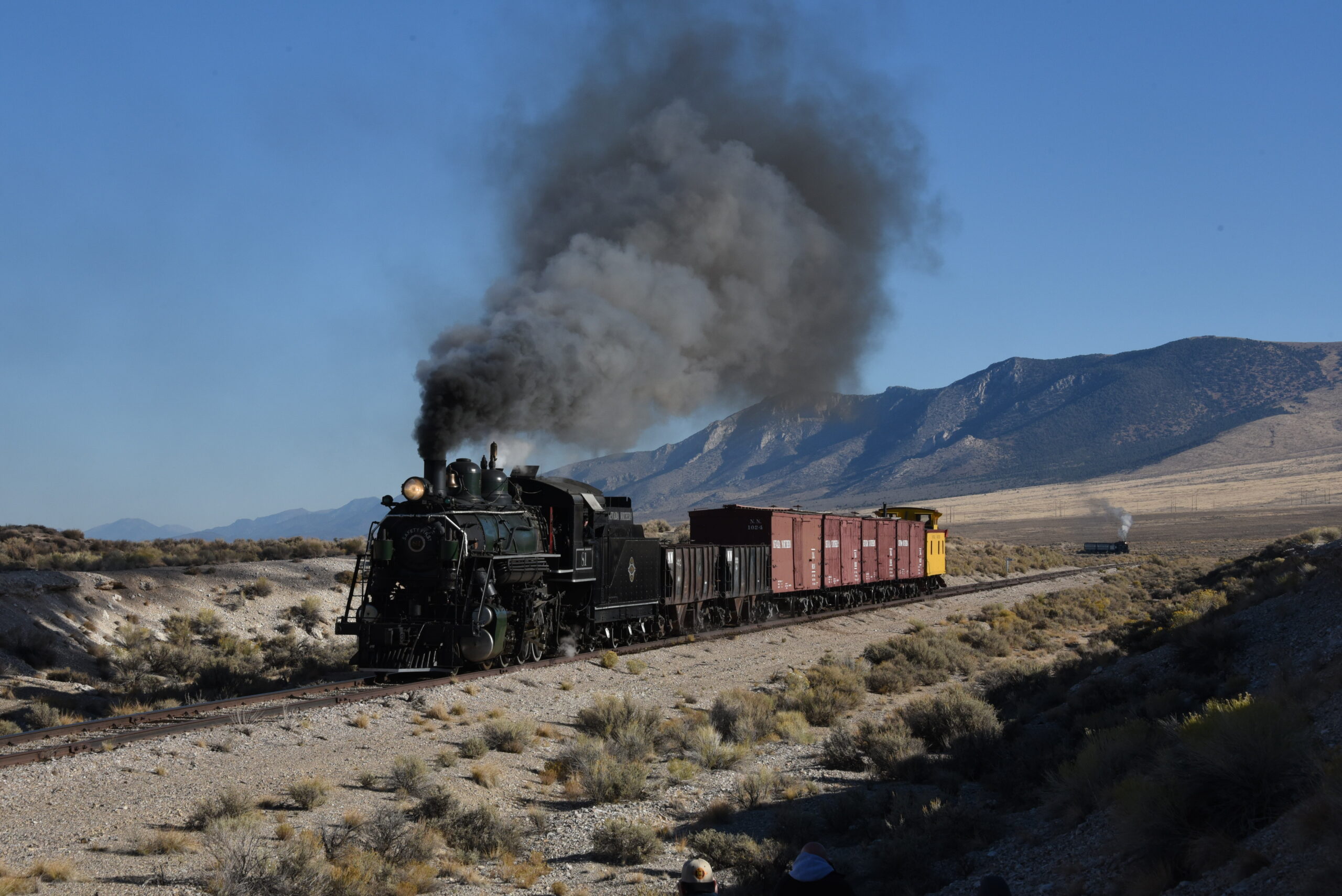 Steam locomotive at speed with freight train while another train waits in the distance.