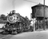 Steam locomotive parked with its nose in a street crossing taking water at a wood water tower