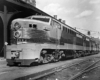 Streamlined, bullet-nosed diesel locomotive sits aside train station canopy