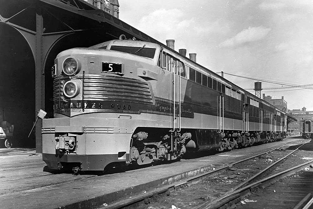 Streamlined, bullet-nosed diesel locomotive sits aside train station canopy
