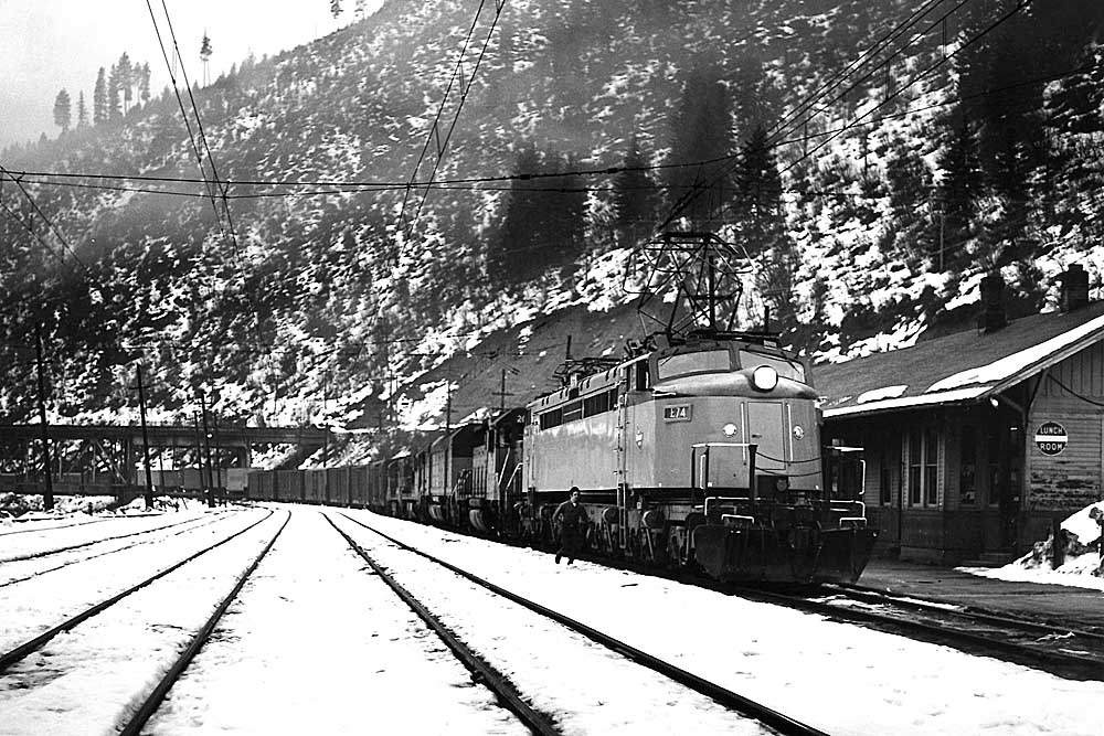 Streamlined electric locomotive leads freight train with four diesels through mountain valley in snow