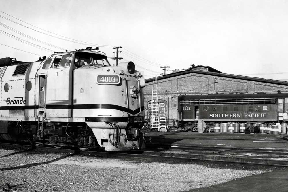 Streamlined diesel locomotive with high cab sits outside brick shop building