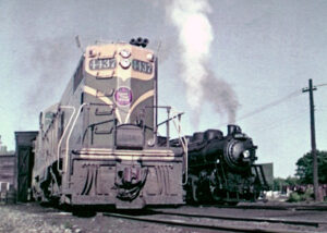 Color photo of road-switcher diesel locomotive and steam locomotive beside each other in yard