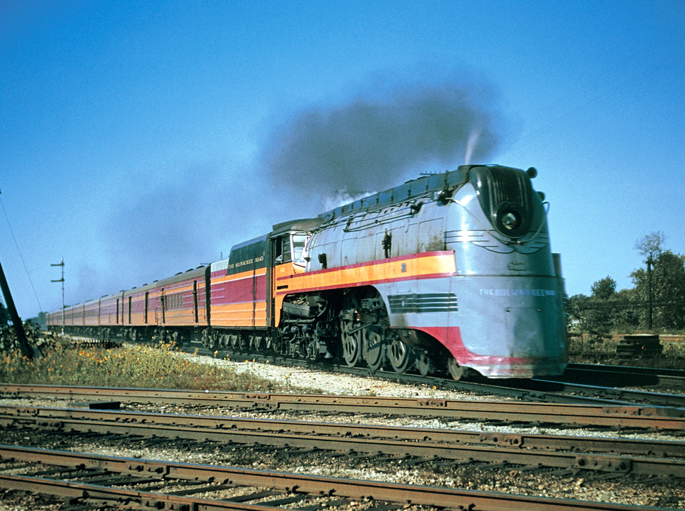 Color three-quarter-angle photo of streamlined steam locomotive with passenger train