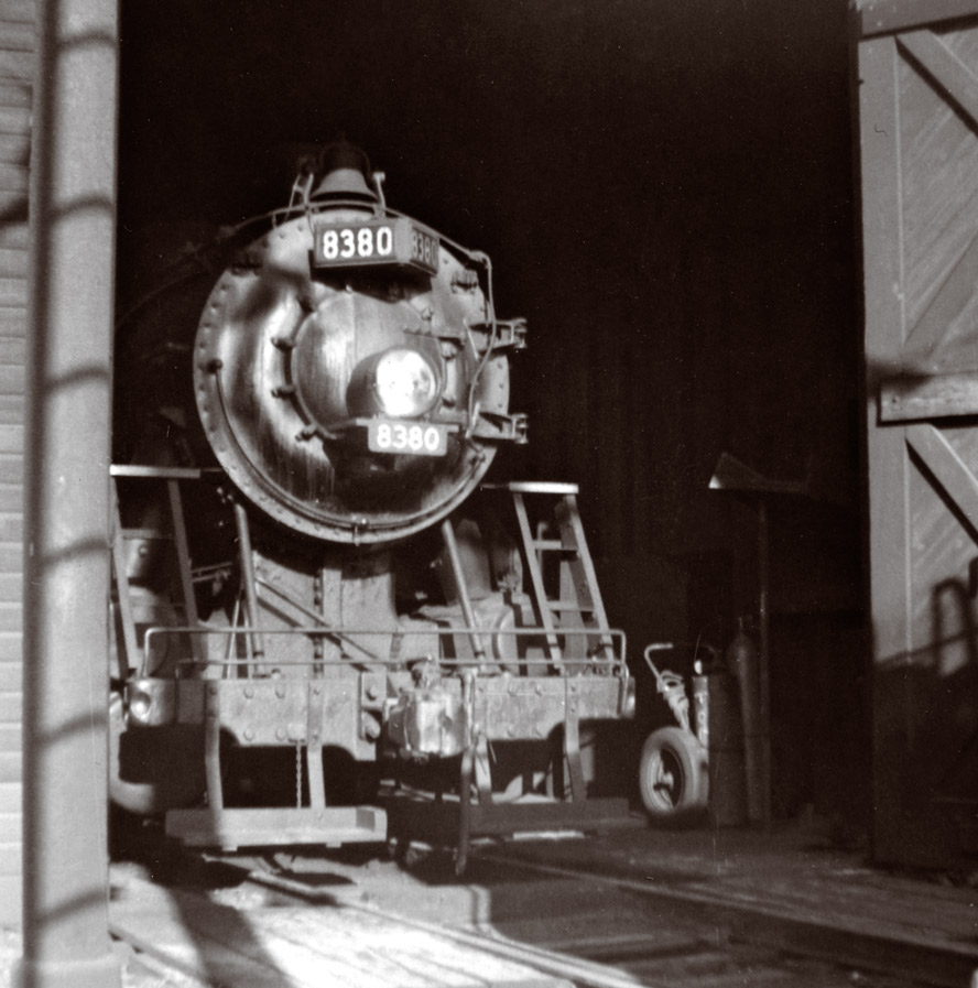 Black-and-white photo of front end of steam locomotive emerging from enginehouse doorway