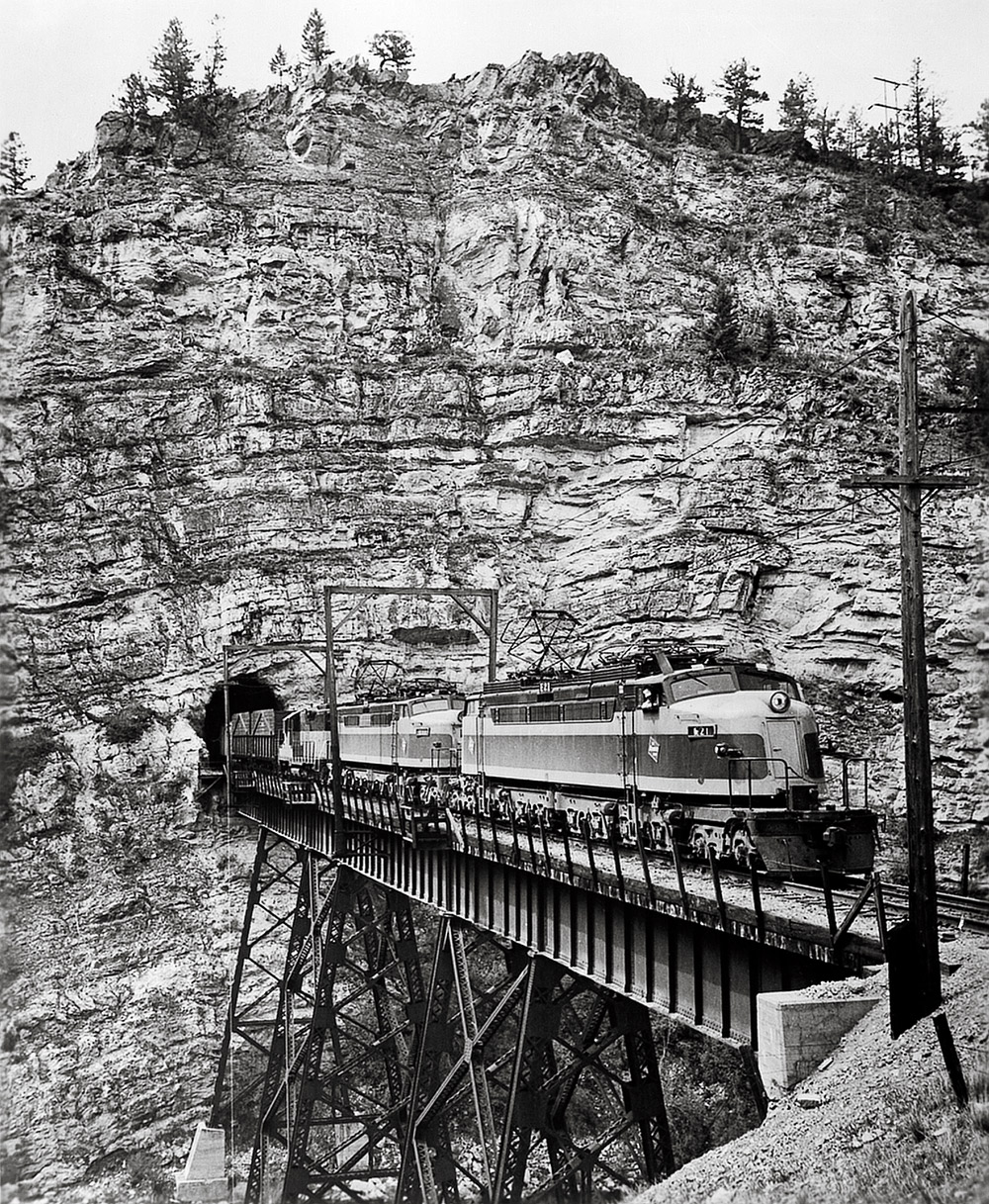 Black-and-white three-quarter-angle photo of streamlined electric locomotives emerging from tunnel and crossing trestle in mountains