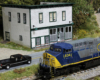 A gray, blue, and yellow modern diesel rolls past a two-story white clapboard country store
