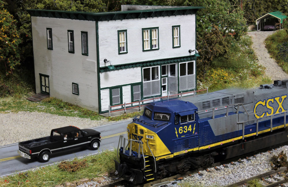 A modern gray, blue and yellow diesel drives past a two-story white shingled country store