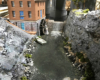 A steam engine pulls a train over a stone arch bridge above a river with a brick mill powered by a water wheel