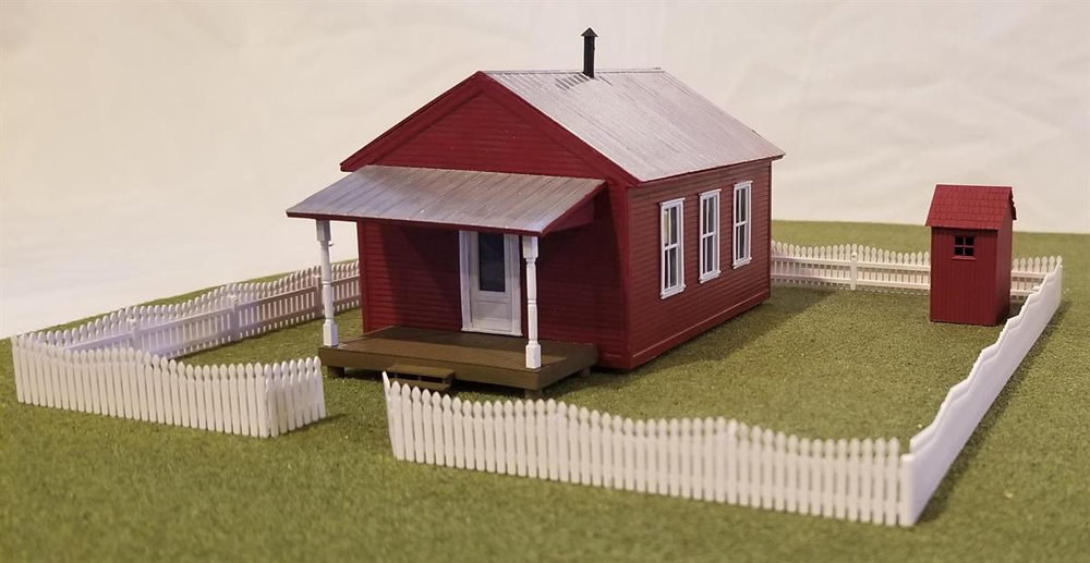 Tichy Train Group HO scale one-room schoolhouse with picket fence and outhouse on scenicked base.