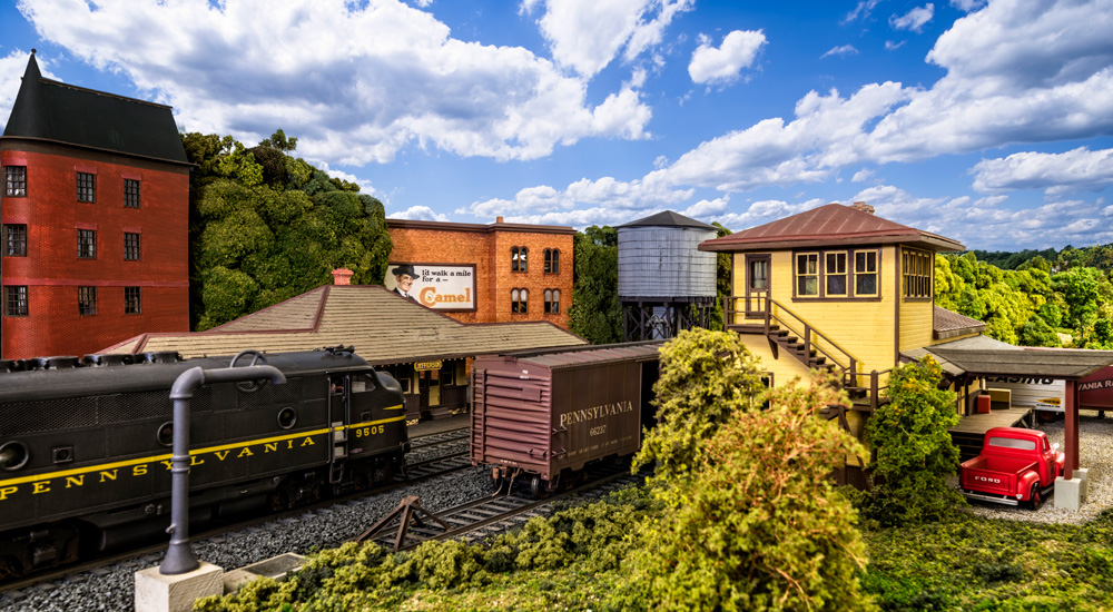 A dark green Pennsylvania Railroad F unit passes a wooden station and a switch tower in a suburban scene