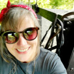 woman in black shirt smiles inside steam locomotive cab; woman in black mask and black shirt inside steam locomotive cab 