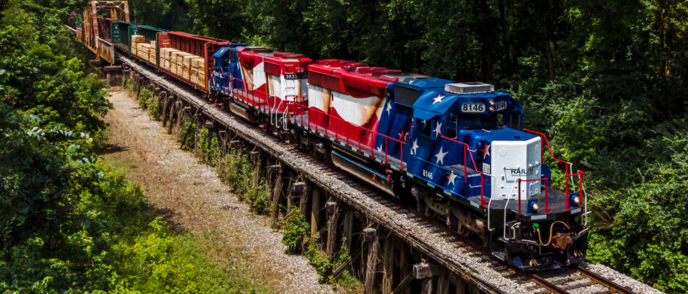 Red, white, and blue locomotives pulling train across low bridge
