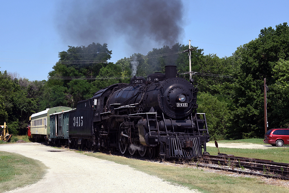 Steam locomotive with passenger car and gondola for riders in field with trees in background.