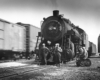 Five men stand in front of steam locomotive while freight passes on adjacent track.