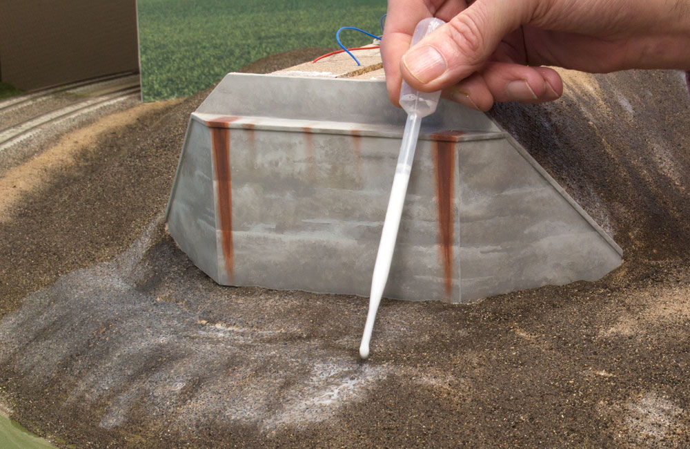 A hand uses a dropper to drizzle scenic cement into ground cover on a train layout