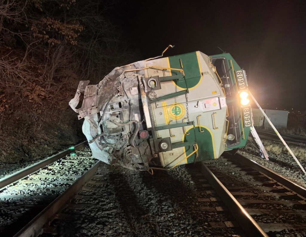 Green, gold, and white locomotive on its side in darkness, headlight still illuminated