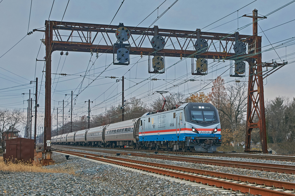 Passenger train with electric locomotive passes under signal bridge.