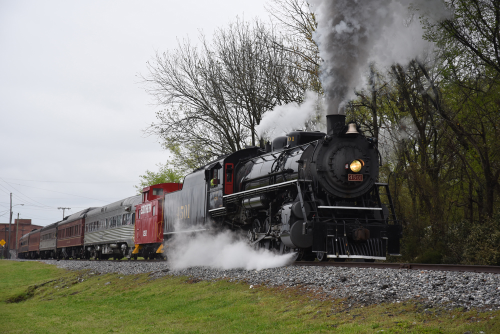 Steam locomotive pulling excursion train with caboose and passenger cars next to line of trees.