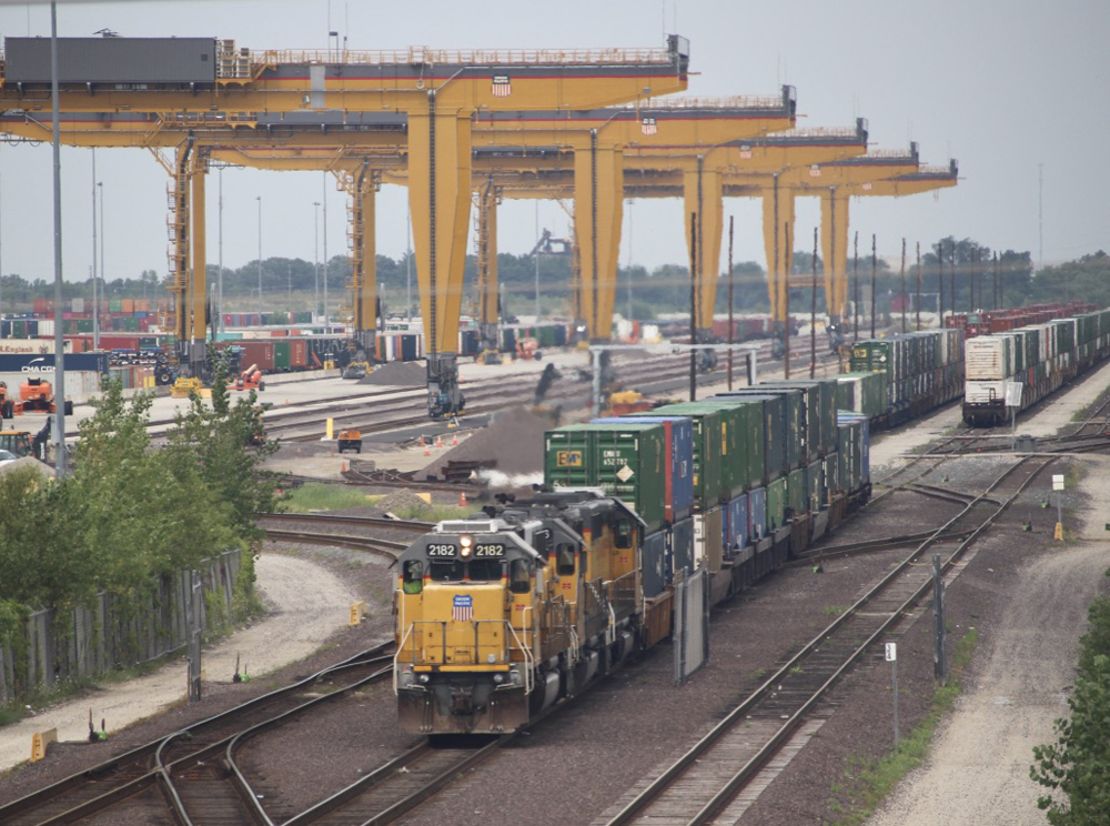 Yellow locomotives moving double-stack cars with container crains in background