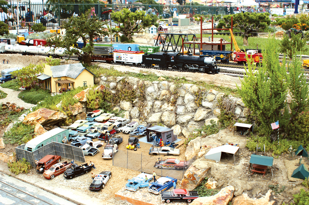 Steam locomotive passes near an auto junk yard on a garden railway