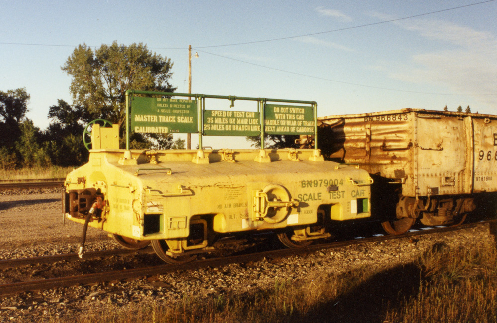 A yellow scale test car with green signs on the top is pulled behind a train.