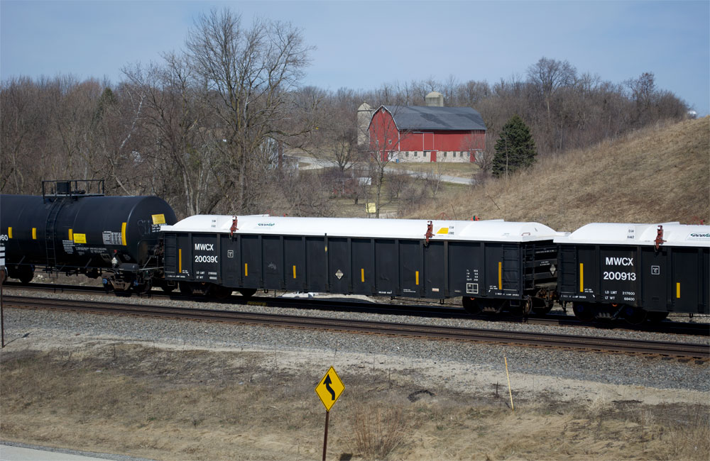 A black gondola with a white Fiberglas cover is seen in a train in front of a red barn