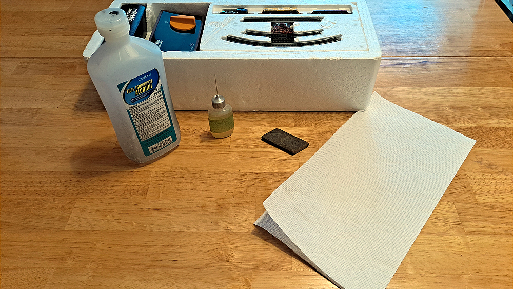 70 percent isopropyl alcohol, plastic-compatible oil in needle-nosed bottle, Bright Boy track cleaner bar, paper towel in front of Styrofoam trainset box insert.