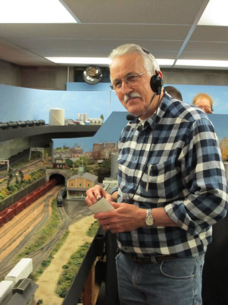 Man with plaid shirt stands next to a model train layout